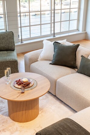 Un salon moderne avec une table basse ronde, un canapé et une vue sur une étendue d’eau grâce à de grandes fenêtres.