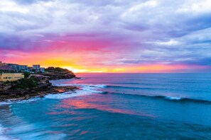 Un paysage côtier avec des bâtiments situés sur un promontoire rocheux, des vagues de l’océan et un coucher de soleil.