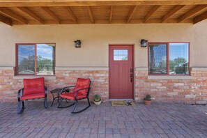 A covered patio with two rocking chairs, a red door, and a brick wall. - Quite 1-bedroom house with mountain views in Oro Valley. (Oro Valley)