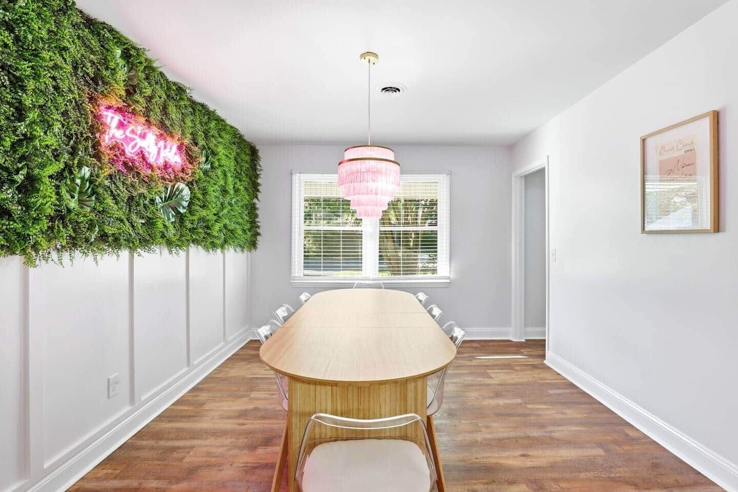 A dining room with a long table, a chandelier, a green wall with a neon sign, and a window with blinds.