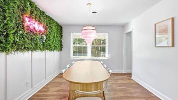 A dining room with a long table, a chandelier, a green wall with a neon sign, and a window with blinds.