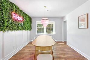 Un comedor con una larga mesa, un candelabro, una pared verde con un letrero de neón y una ventana con persianas.