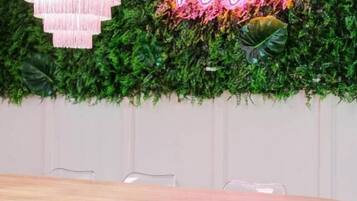 A dining area with a round table, clear chairs, a pink chandelier, and a neon sign on a green leafy wall.