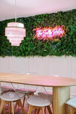 Een eetruimte met een ronde tafel, transparante stoelen, een roze kroonluchter en een neonbord op een groene, begroeide muur.