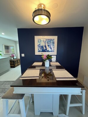 Un coin repas avec une table dressée pour quatre personnes, un vase rempli de fleurs, un tableau encadré au mur et un luminaire suspendu au plafond.