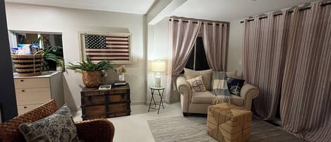 Un salon comprenant un canapé, une table basse, un fauteuil en osier, une petite table avec une lampe, une commode décorative et un drapeau américain accroché au mur.