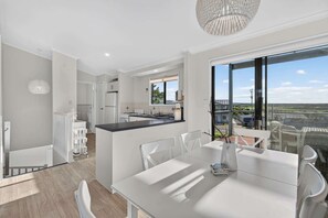 A modern kitchen with a central island, a dining area with a white table and chairs, and a large window offering a view of the countryside. - Sea Break - Middleton Beach Holiday House with Ocean View (Middleton)