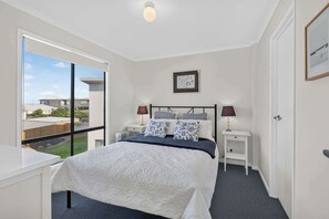 Une chambre à coucher avec un lit, deux tables de chevet avec des lampes, un tableau encadré au mur et une vue sur l’extérieur.
