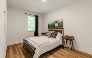 Una camera da letto con un letto di legno, un comodino, una finestra con tende e un'opera d'arte appesa al muro.