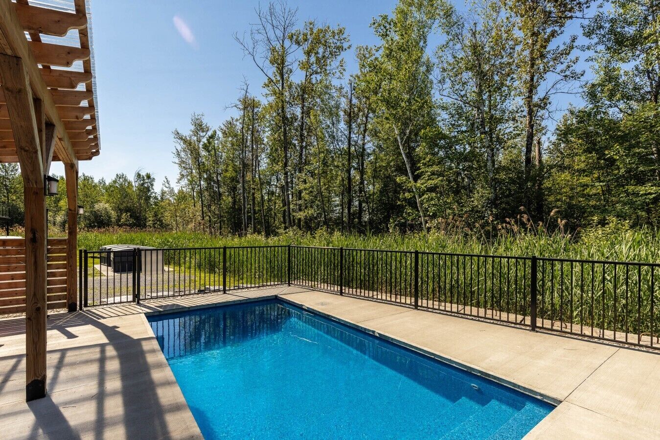 A rectangular swimming pool with a black metal fence, surrounded by a wooden deck and a wooded area.