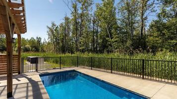 A rectangular swimming pool with a black metal fence, surrounded by a wooden deck and a wooded area.