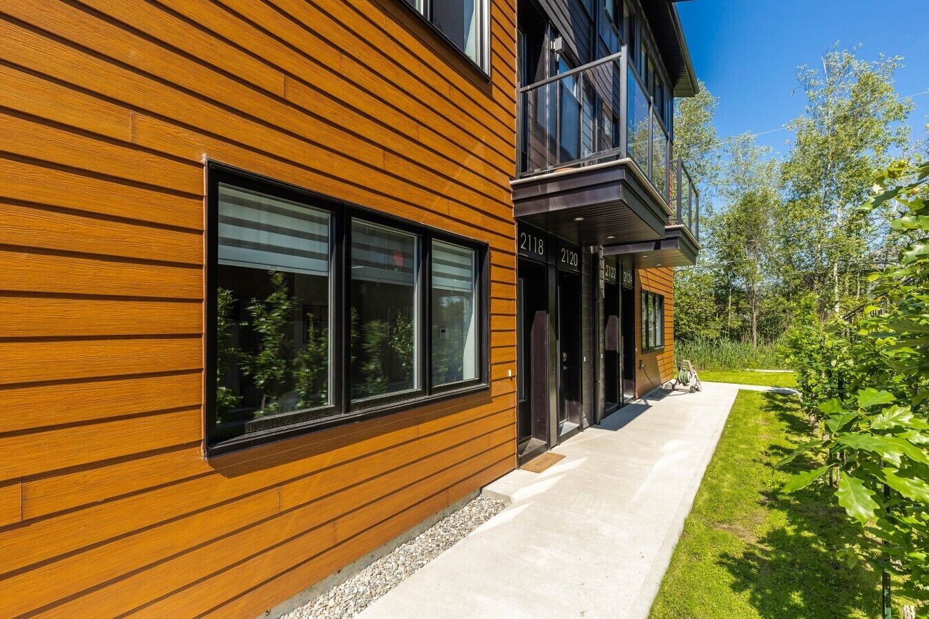 A modern two-story building with horizontal wooden siding, large windows, and a concrete pathway leading to the entrance.