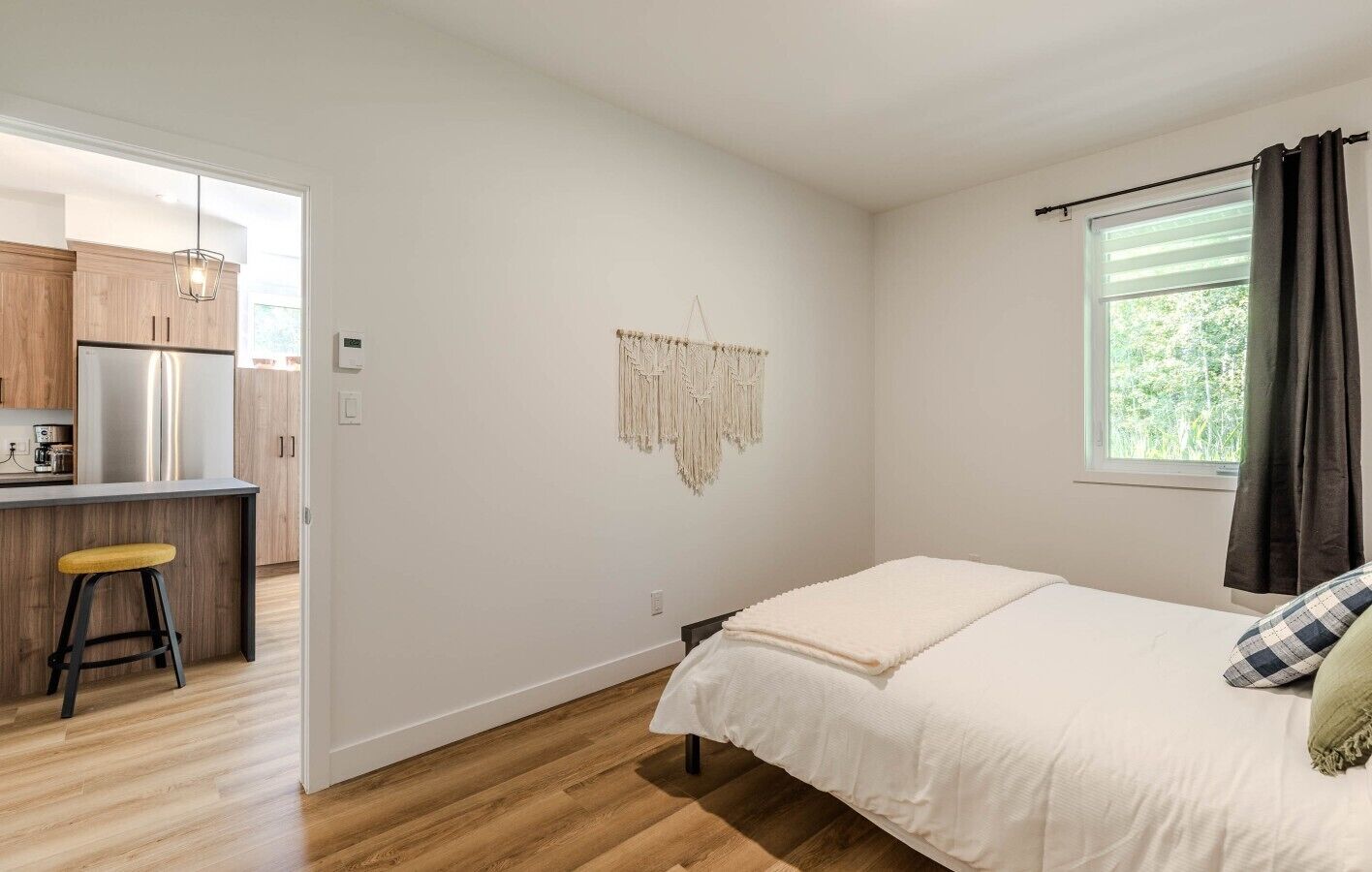 A bedroom with a bed, a window with curtains, a kitchen area with a refrigerator, and a macrame wall hanging.