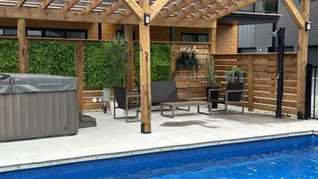 A covered outdoor area with a pool, wooden pergola, lounge chairs, and a hot tub.