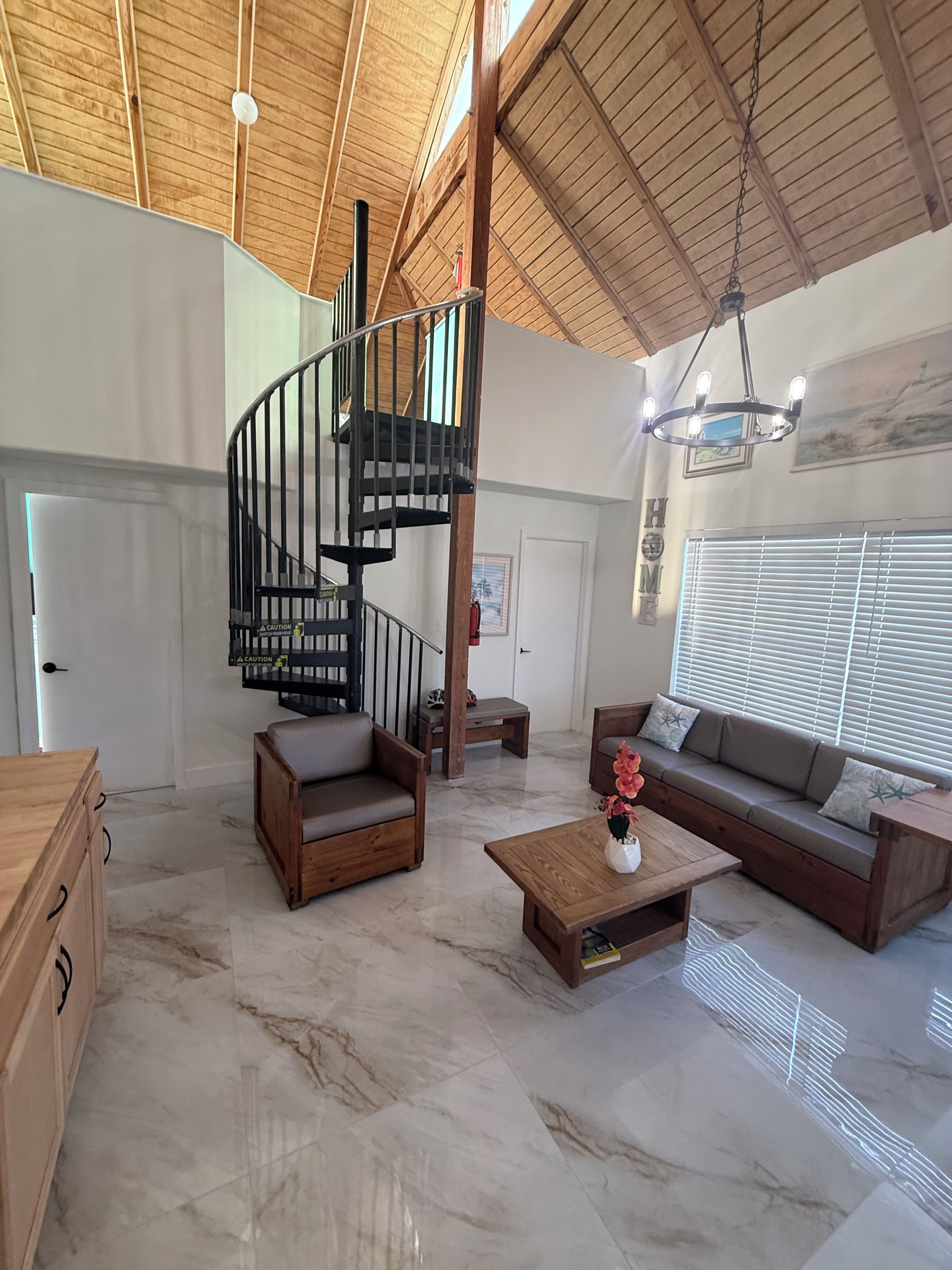 A spacious living area with a wooden staircase, a sofa set, a coffee table, and a large window with blinds.