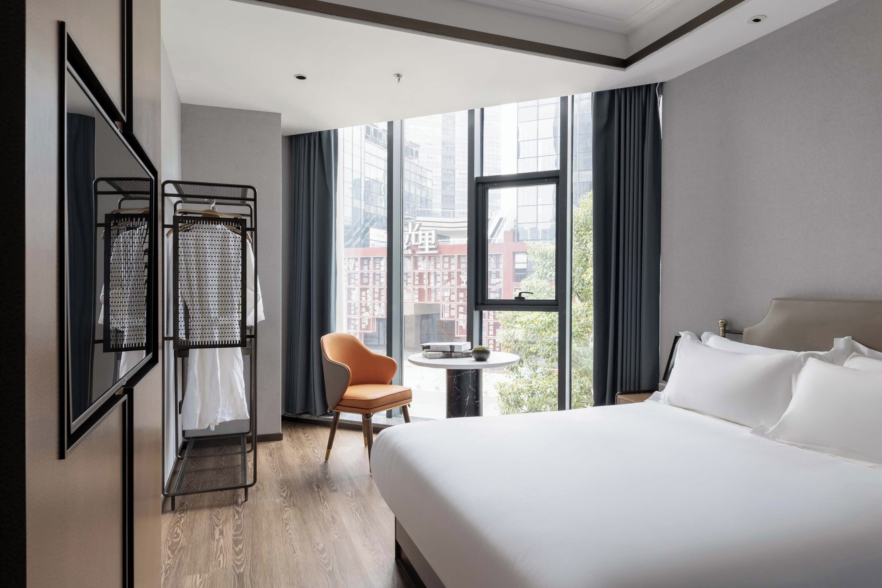 A hotel room with a large bed, a chair, a small table, and a window with a view of buildings.