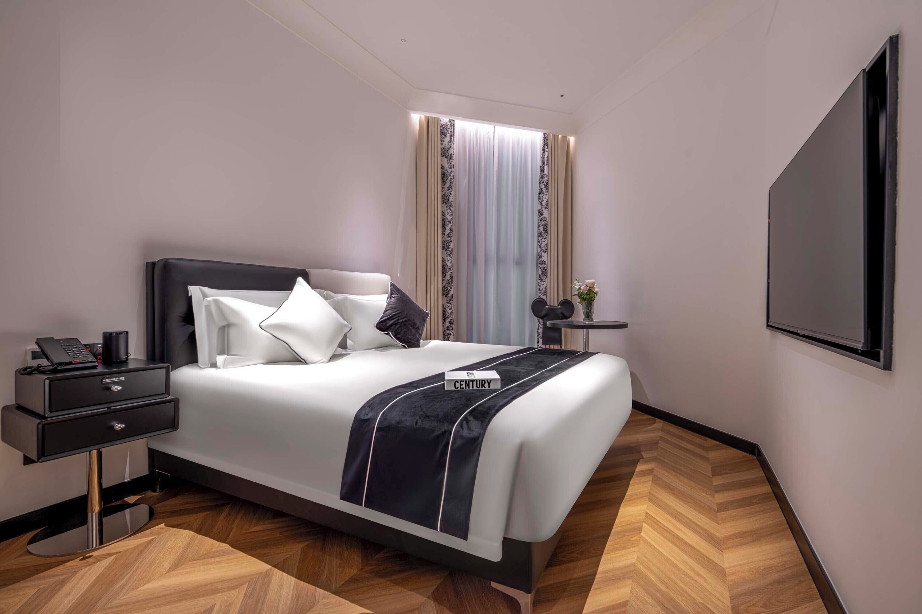 Habitación de hotel moderna con una cama grande, una mesita de noche con un teléfono, una pequeña mesa con un florero y un espejo grande en la pared.