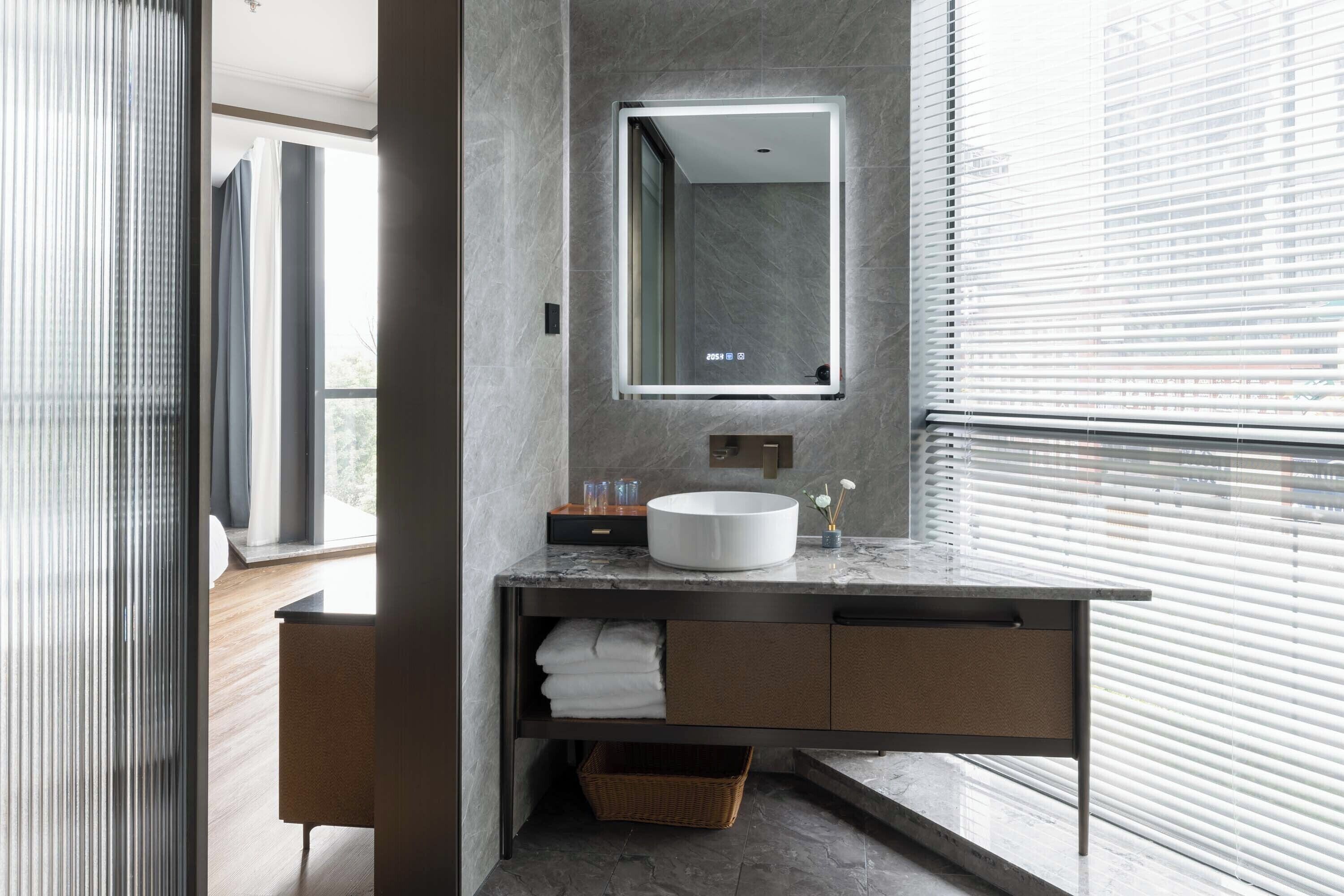 A modern bathroom with a marble countertop, a round sink, a mirror, and a window with blinds.