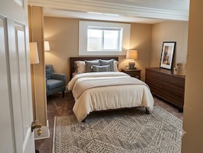 Um quarto com cama, luminárias de cabeceira, uma cadeira, cômoda e janela.
