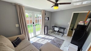Ein Wohnzimmer mit einer Couch, einem Deckenventilator, einem Schreibtisch und Blick durch eine Glastür in einen Garten.