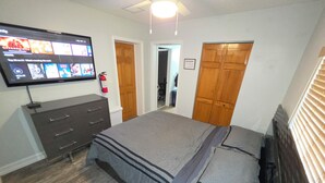 Ein Schlafzimmer mit einem Bett, einem Schminktisch, einem an der Wand befestigten Fernseher und einer Tür zu einem anderen Raum.