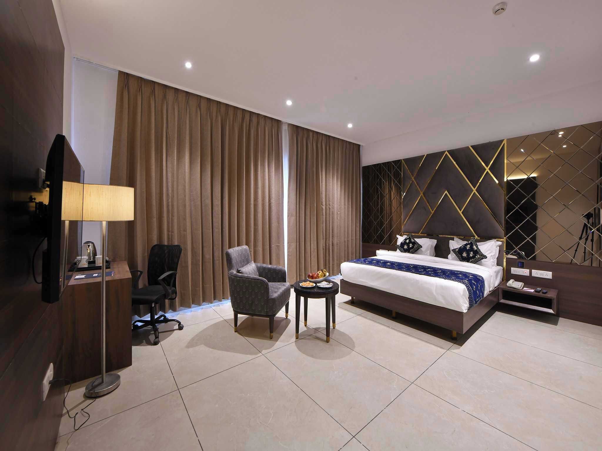 A hotel room with a bed, a desk with a lamp, a chair, a small table with snacks, and a television.