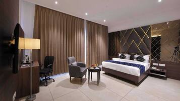 A hotel room with a bed, a desk with a lamp, a chair, a small table with snacks, and a television.
