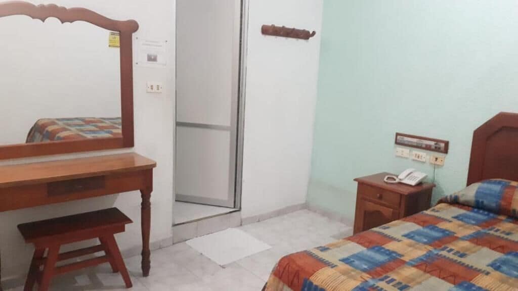 A bedroom with a bed, a wooden desk, a chair, a bedside table with a phone, and a mirror.