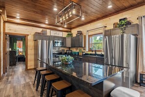 A modern kitchen with stainless steel appliances, a large island with bar stools, wooden ceiling, and a view of the outdoors. - Luxury BRAND NEW 8bdrm Cabin-Pool-Arcades-Gazebo (Sevierville)