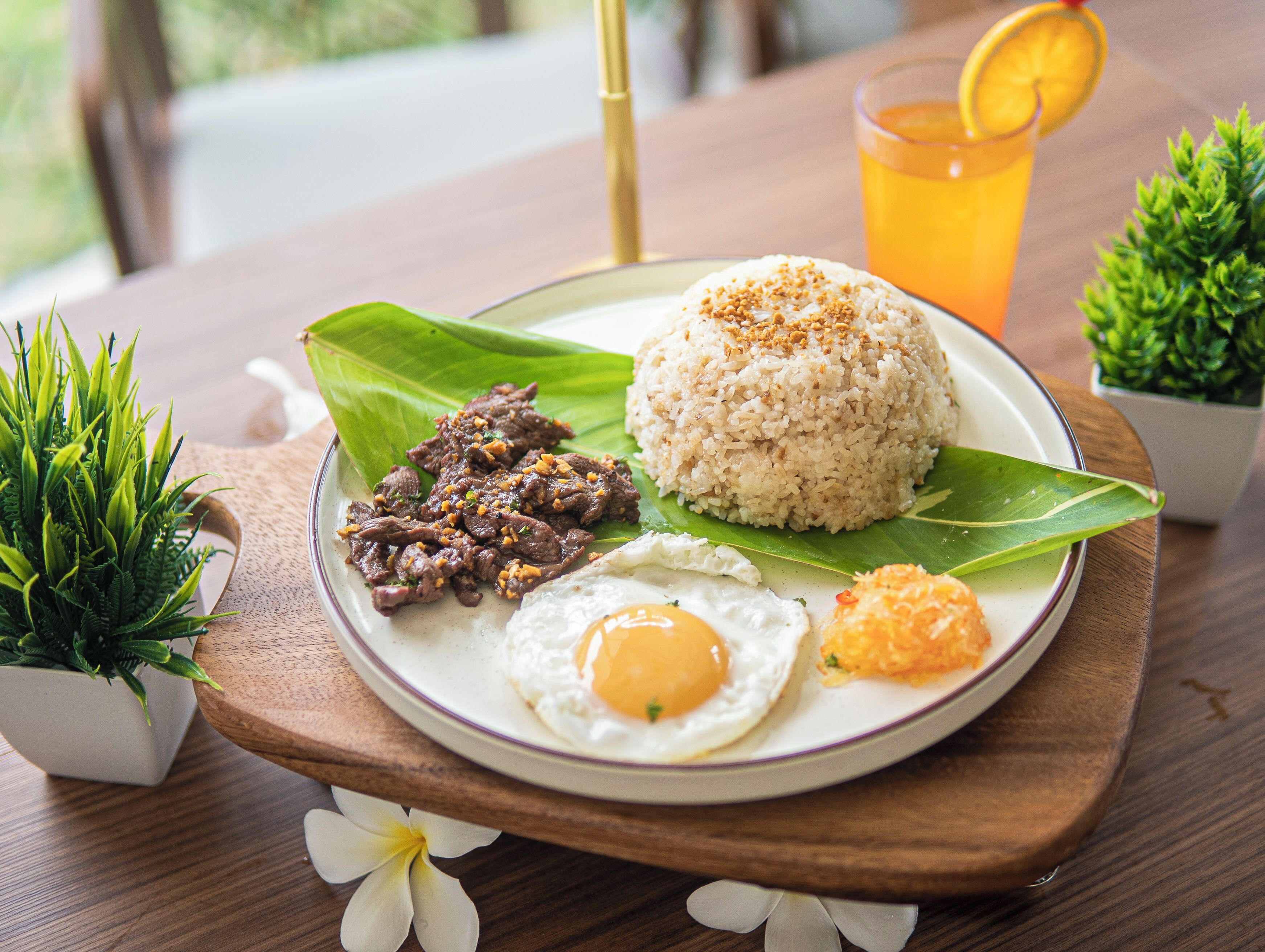 A plate of food with rice, beef, egg, and a side dish, served on a wooden table with a glass of orange drink.
