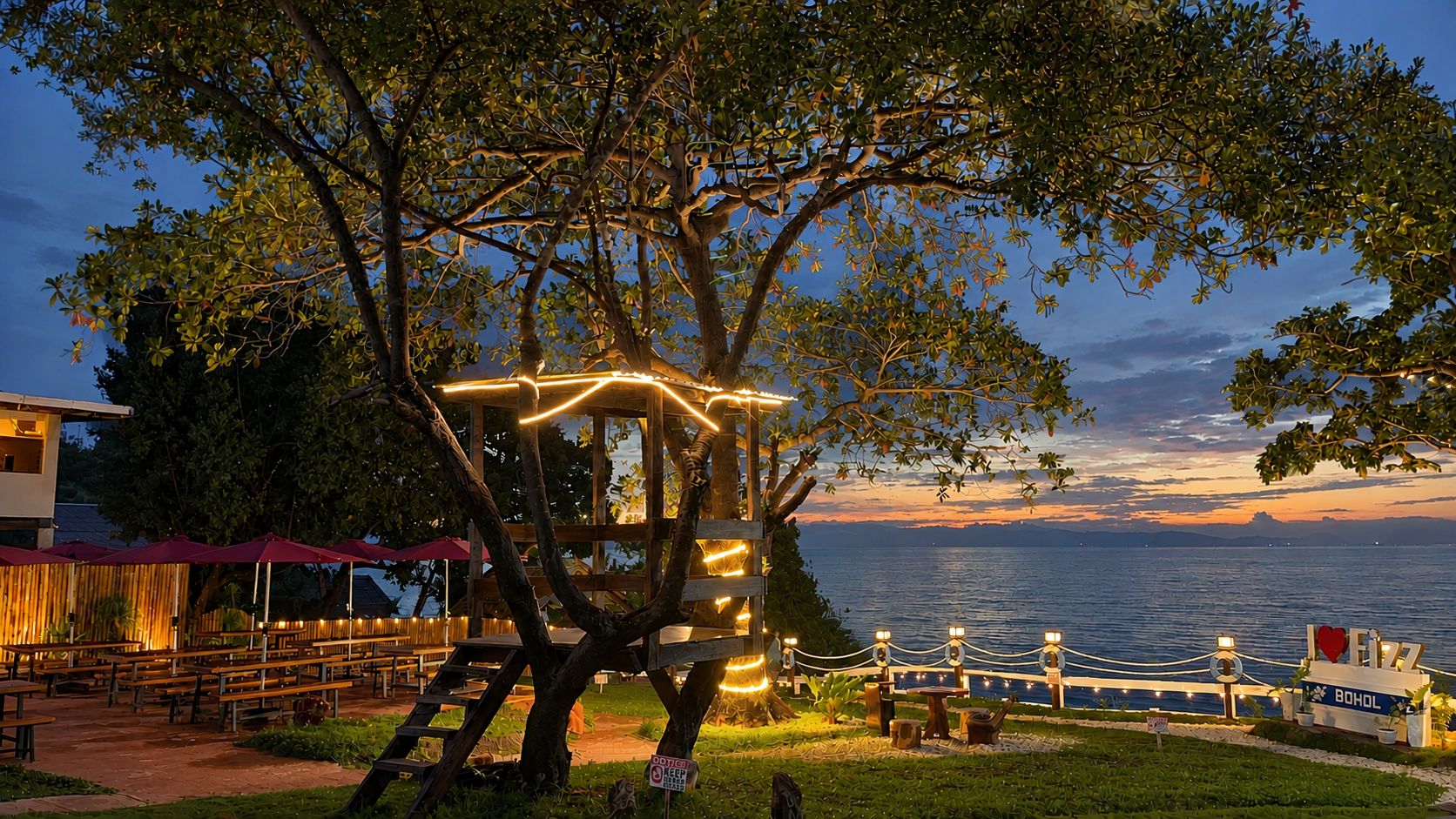 Estructura tipo casa en un árbol con luces colgantes, con vistas a un cuerpo de agua.