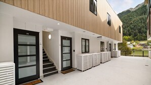 En bygning med moderne arkitektur, med en trappe op til en dør med skiltet '29B', et overdækket udendørs område med hvide bænke, og et skovklædt bjerg i baggrunden.