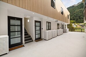 Ein Gebäude mit modernem Design, das eine Treppe zu einer Tür mit der Aufschrift '29B', einen überdachten Außenbereich mit weißen Bänken und einen Blick auf einen bewaldeten Berg im Hintergrund zeigt.