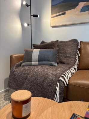 Ein braunes Ledersofa mit einem grau-weiß gestreiften Kissen, einer grauen Decke mit weißen Fransen und einem kleinen Holztisch mit einem mit 'UPLIFT' beschrifteten Glas in einem modernen Raum mit einem Bild an der Wand.