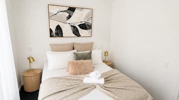 A neatly arranged bedroom with a bed, pillows, a blanket, a wall-mounted light, and a framed abstract artwork.