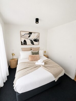 A neatly arranged bedroom with a bed, pillows, a blanket, a wall-mounted light, and a framed abstract artwork. - Lake and Mountain Views (Queenstown)