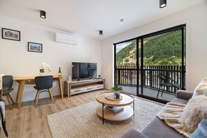 Ein modernes Wohnzimmer mit Fernseher, Sitzgelegenheiten, einem Essbereich und einem Balkon mit Blick auf ein bewaldetes Gebiet.
