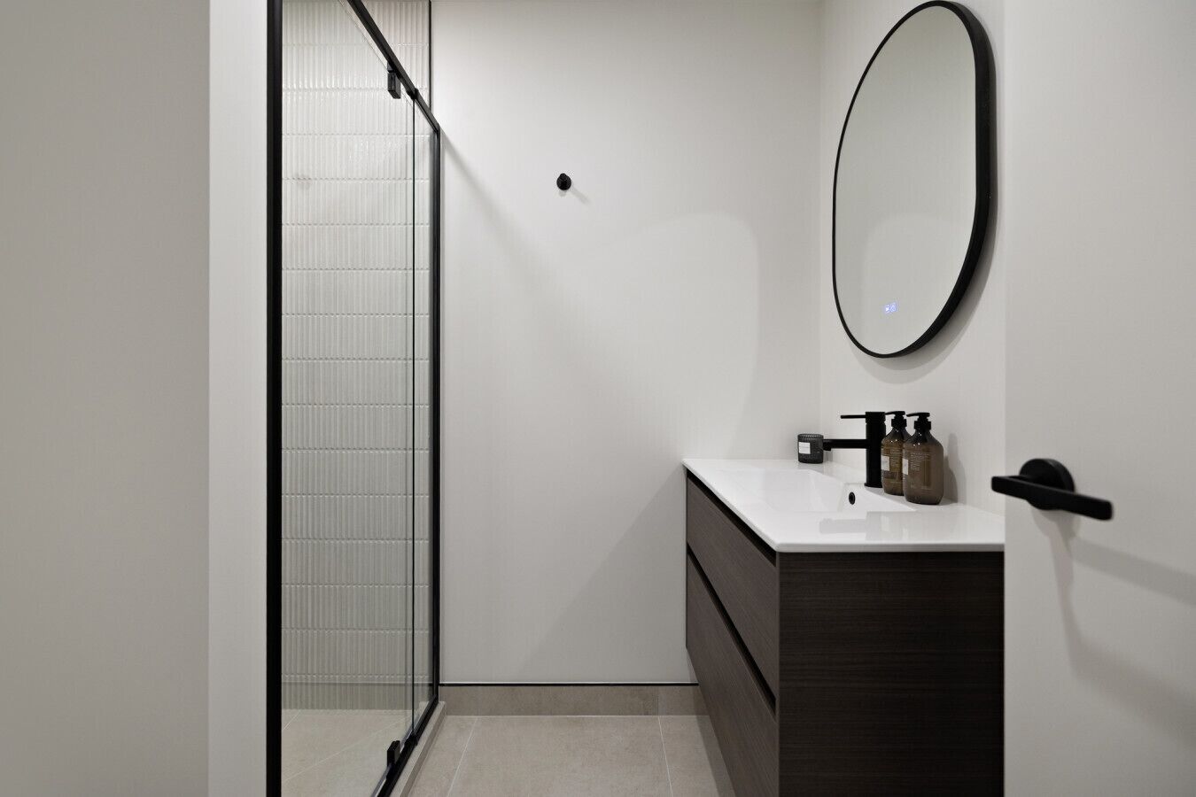 A modern bathroom with a glass shower enclosure, a white sink with dark cabinetry, and a round mirror on the wall.