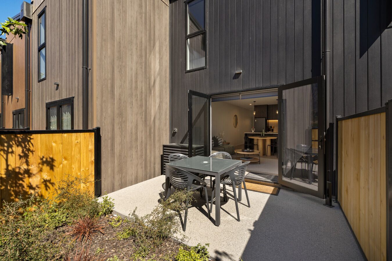 En moderne bygning med en patio, et bord, stole, et træhegn og udsigt ind i interiøret.