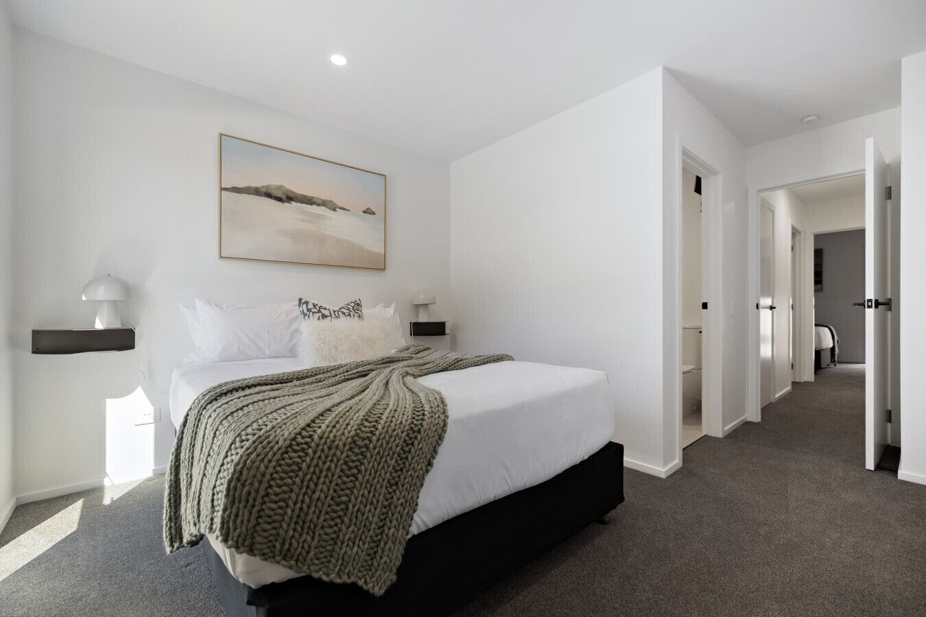 A modern bedroom with a large bed, a painting above the bed, and a hallway leading to other rooms.