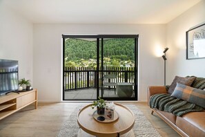 A living room with a large sliding glass door leading to a balcony, a television on a wooden stand, a round wooden coffee table with a plant, and a brown leather sofa with a blanket. - Lake and Mountain Views (Queenstown)
