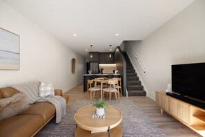 Ein modernes Wohnzimmer mit einer Couch, einem Couchtisch und einem Fernseher. Es gibt eine Küchenzeile mit einem Esstisch und Stühlen. Im Hintergrund ist eine Treppe zu sehen.