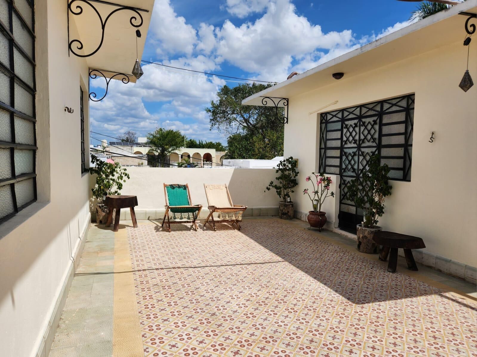 A covered outdoor patio with patterned flooring, a seating area with chairs, and potted plants.