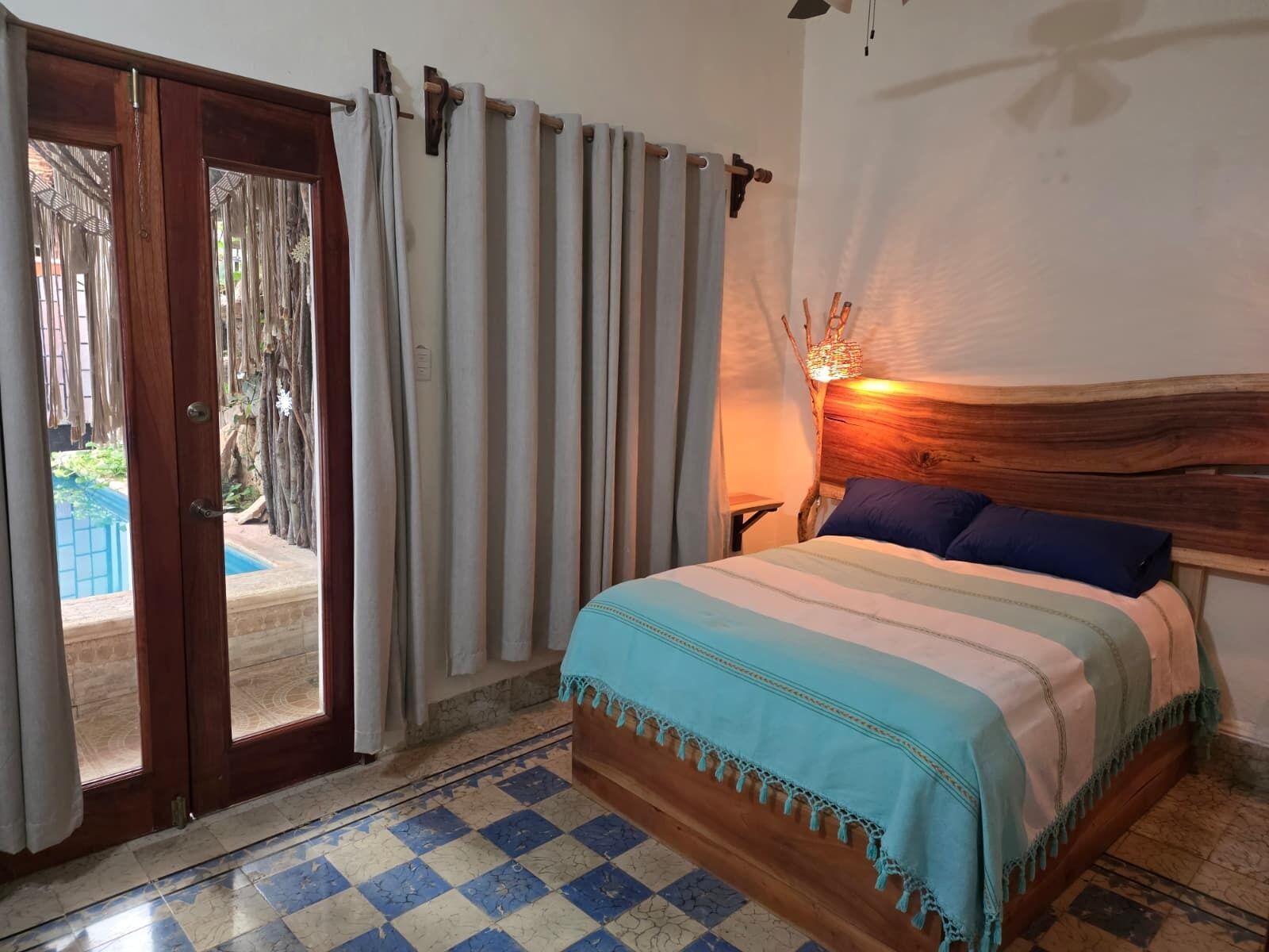 Une chambre à coucher avec un lit, des rideaux, une tête de lit en bois, une table de chevet et une vue sur une piscine extérieure.