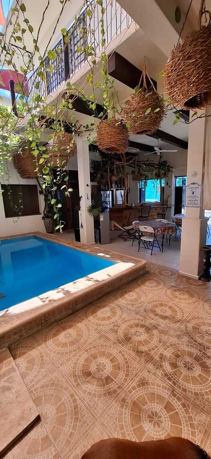 An indoor swimming pool with a tiled floor, surrounded by a dining area with chairs and tables, and hanging baskets with plants.