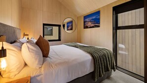 Un dormitorio con cama, almohadas, una lámpara de noche, una ventana y un cuadro de la ciudad.