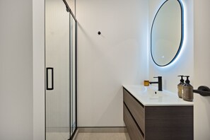 Un bany modern amb plat de dutxa de vidre, un lavabo blanc i un moble sota el lavabo fosc, i un mirall gran amb il·luminació circular.