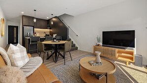 A modern living room with a sofa, coffee table, and a TV. There's a dining area with chairs and a table. A kitchen is visible in the background. - Prime Location with mountain views (Queenstown)