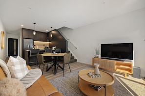 Ein modernes Wohnzimmer mit Sofa, Couchtisch und Fernseher. Es gibt einen Essbereich mit Stühlen und Tisch. Im Hintergrund ist eine Küche zu sehen.