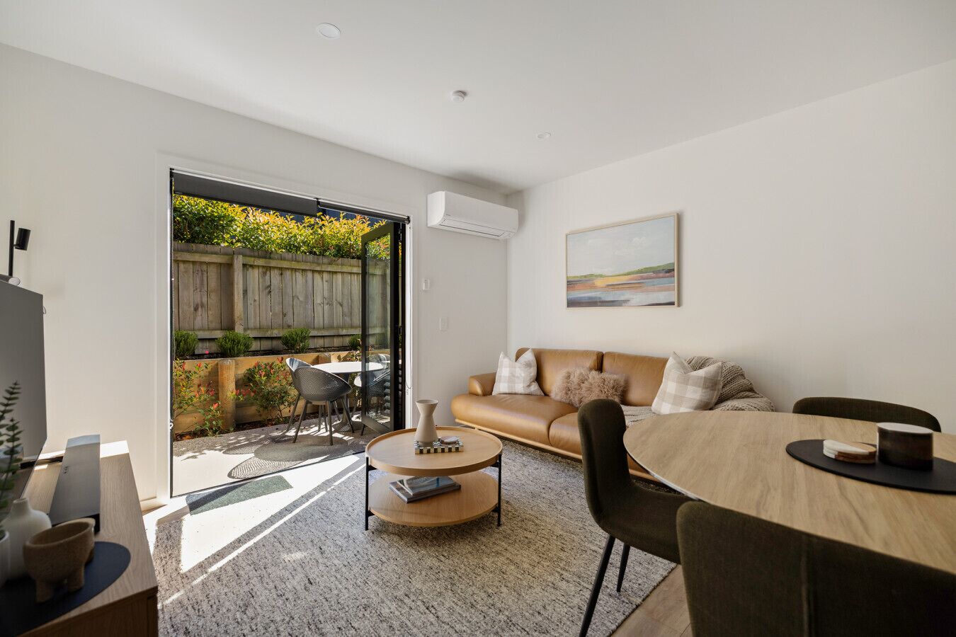 A modern living room with a sofa, coffee table, and dining table. There is a sliding glass door leading to a patio with a wooden fence and plants.